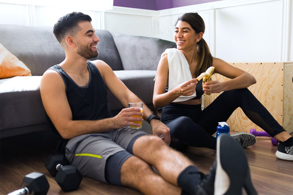 A man drinking water and a woman eating a banana while sitting on the living room floor, getting ready for a workout.