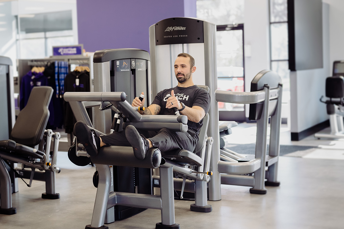 A man in the starting position on a leg curl machine in a gym setting.