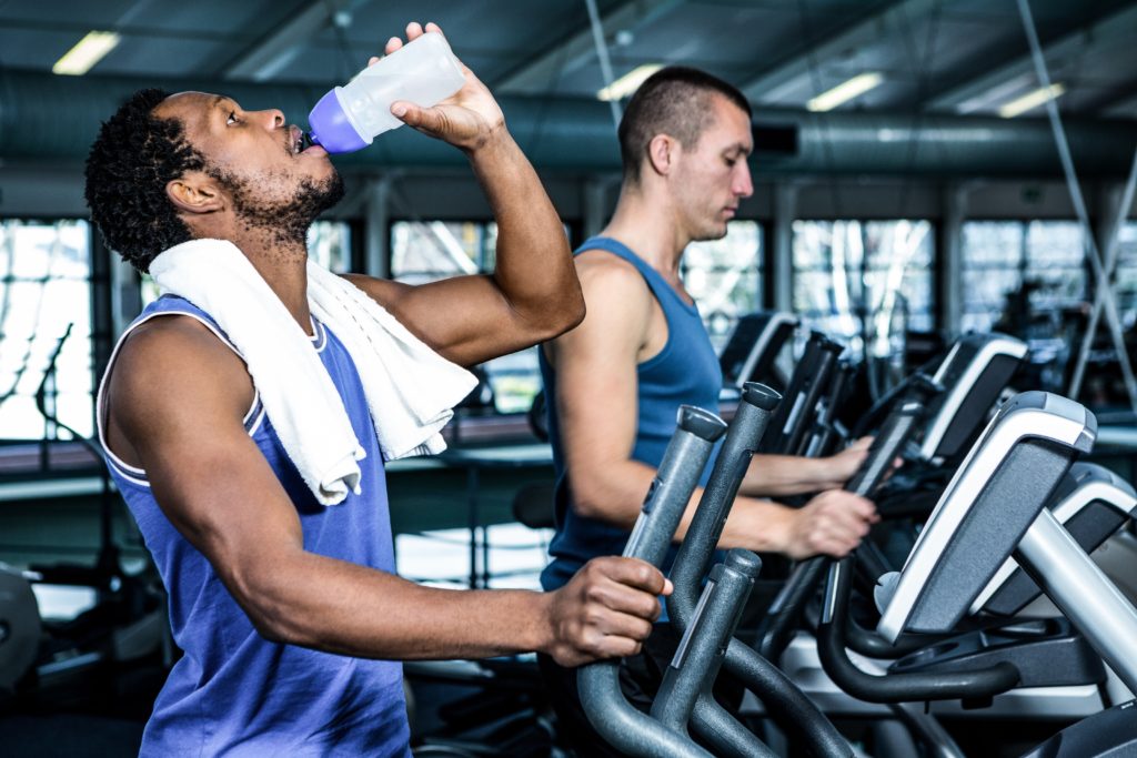 Man drinking water while on a gym elliptical machine, with another man on an elliptical next to him.