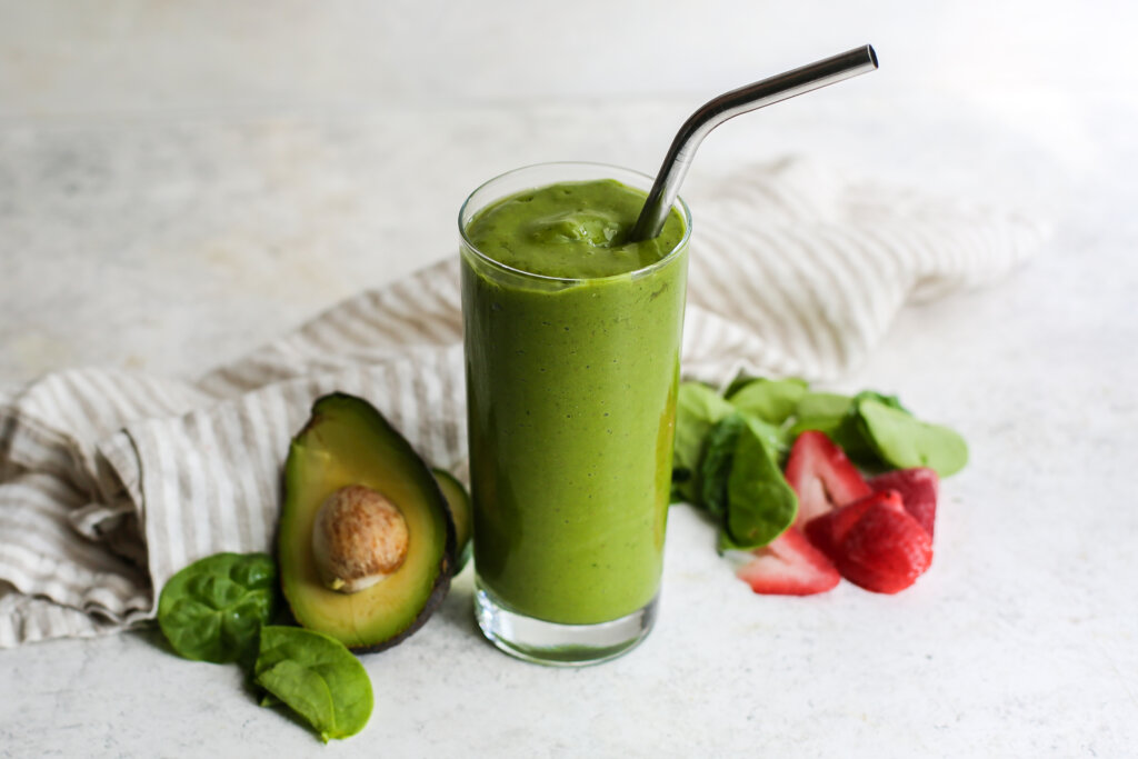 Glass of green smoothie sitting next to half an avocado and sliced strawberries.