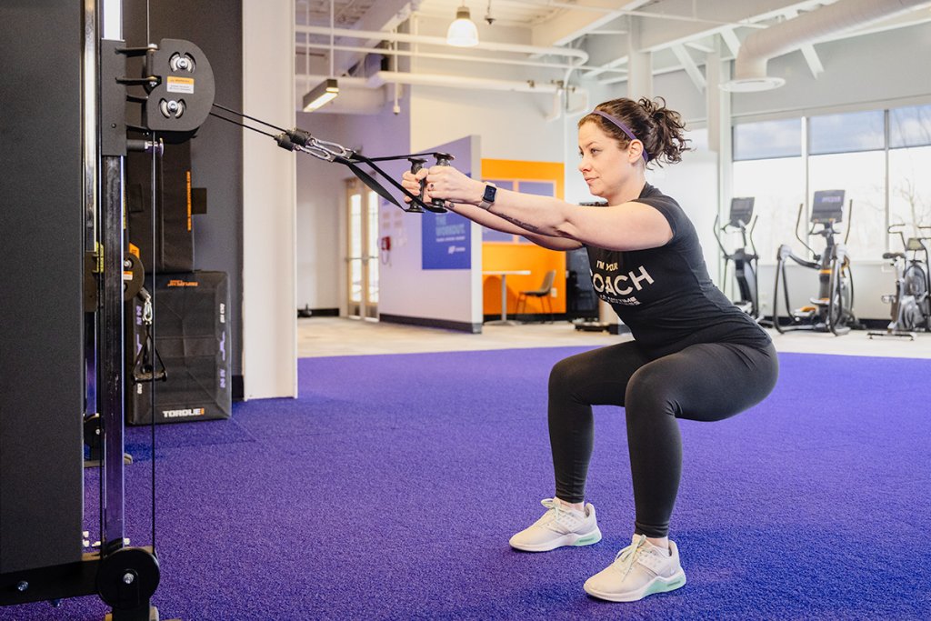 Coach Heather demonstrating a squat to row with cables in a gym setting.