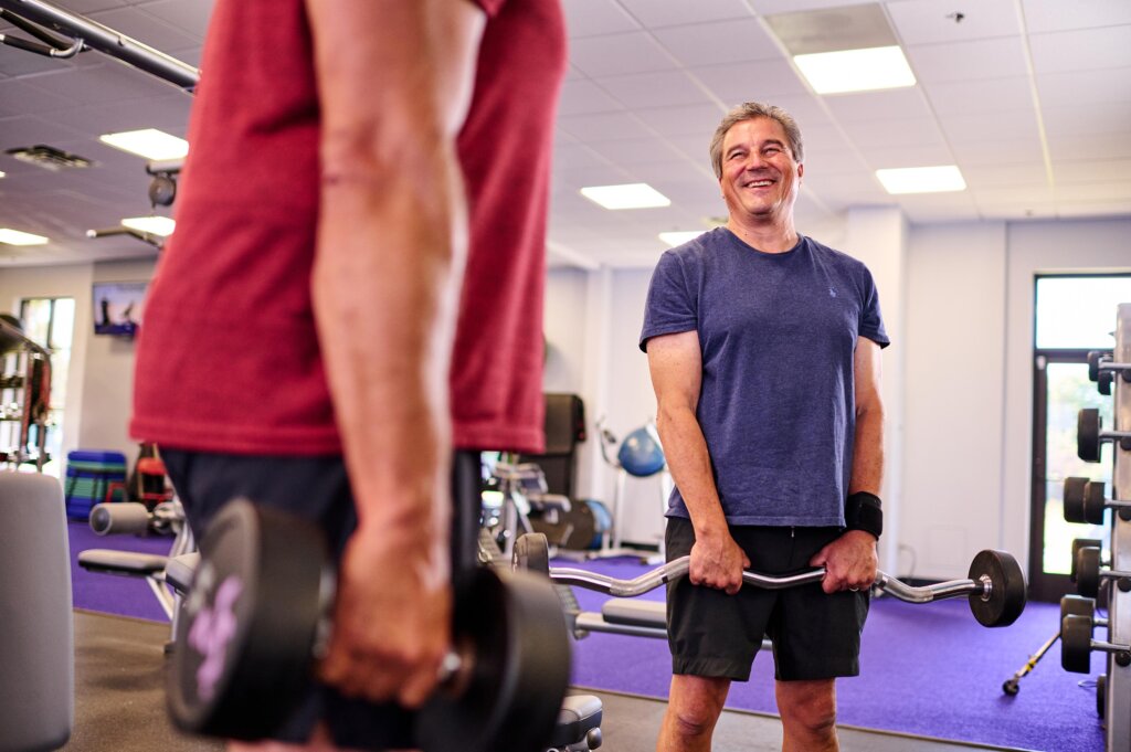 Middle-aged man holds a barbell and smiles at his gym companion.