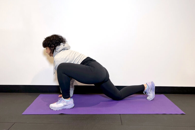 Coach on a yoga mat with her right leg back straight and her left leg bent with foot a planted on the ground.