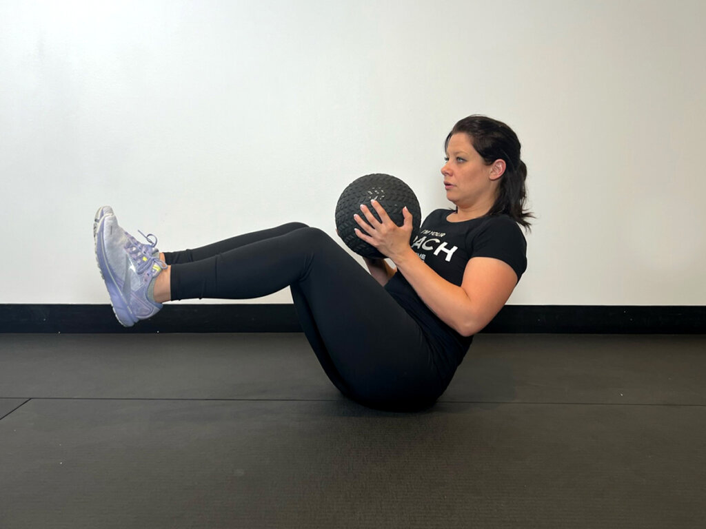 Coach seated with knees in the air, holding medicine ball with both hands at chest.