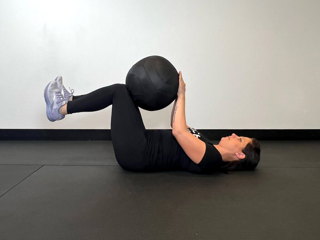 Coach lying on back holding a medicine ball between hands and knees.