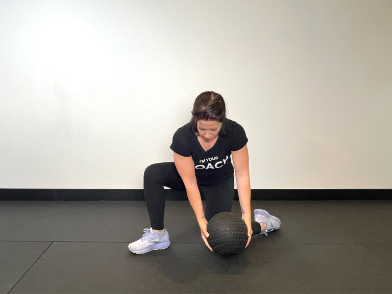 Coach in lunge position with left knee on the ground, torso twisted to the left with outstretched arms holding medicine ball down at ground level.