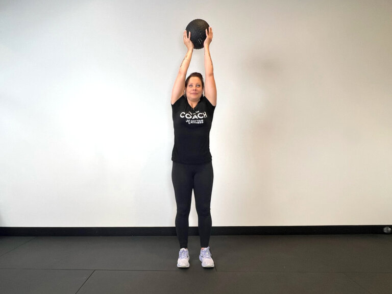 Coach standing, holding medicine ball above head with both hands.