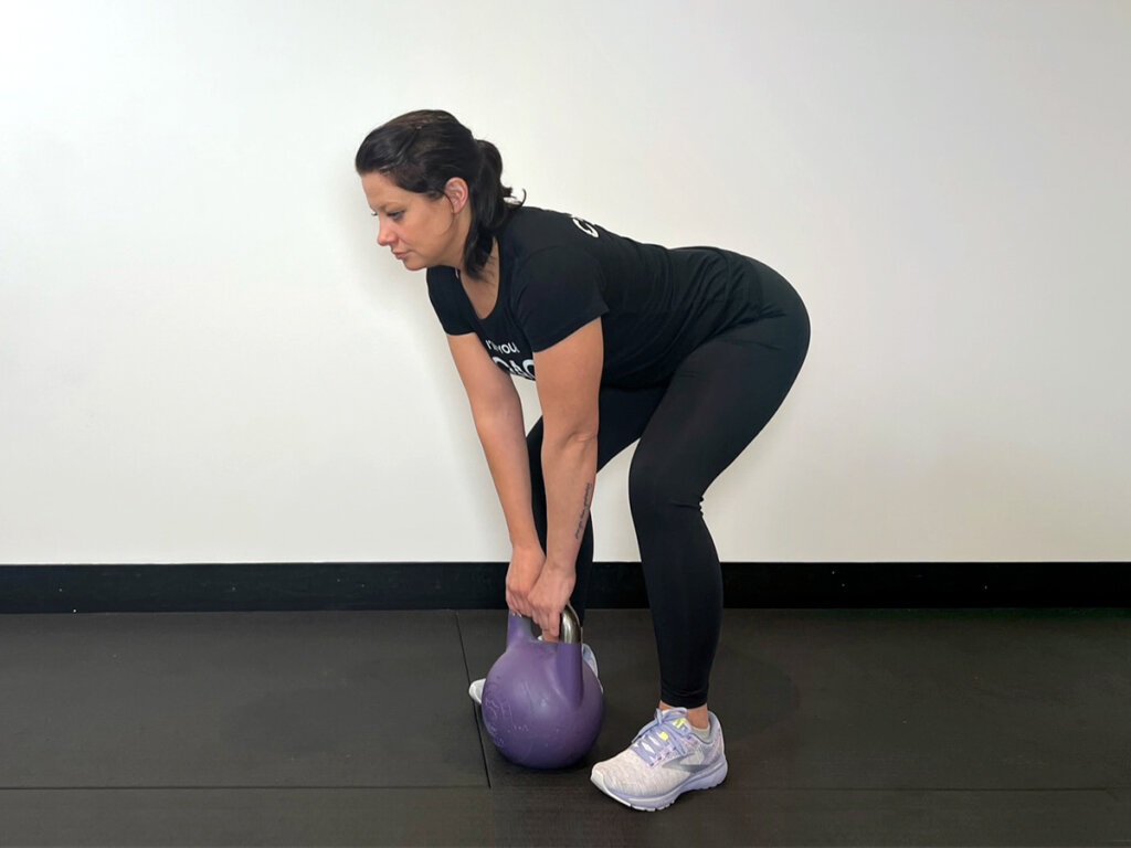 Coach holding kettlebell with both hands between legs, slightly squatting.