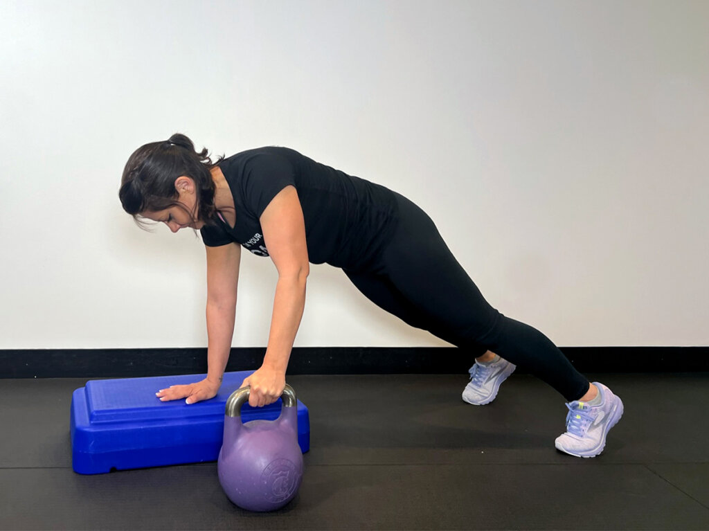 Coach in a horizontal plank position with one hand resting on exercise block and the other hand holding a kettlebell that’s resting on the ground.