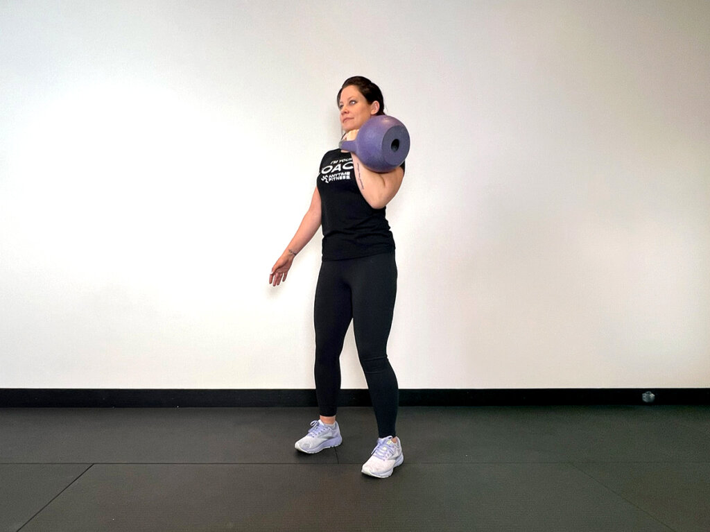 Coach holding kettlebell in one hand at shoulder level in a rack position.