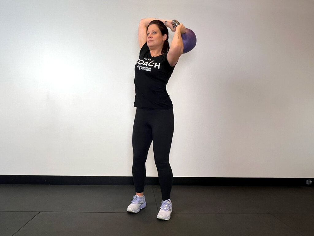 Coach standing straight, holding kettlebell with both hands and moving it in a “halo” motion around head.