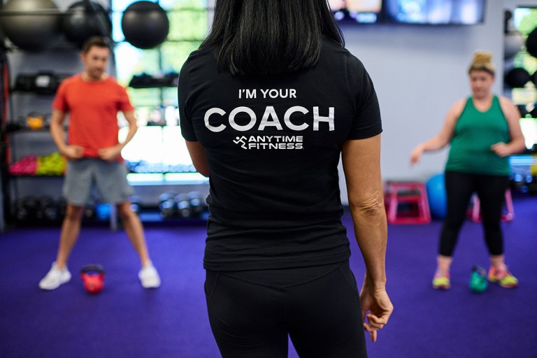 An Anytime Fitness personal trainer wearing a T-shirt that says “I’m Your Coach. Anytime Fitness” is facing two gym members during a training session.