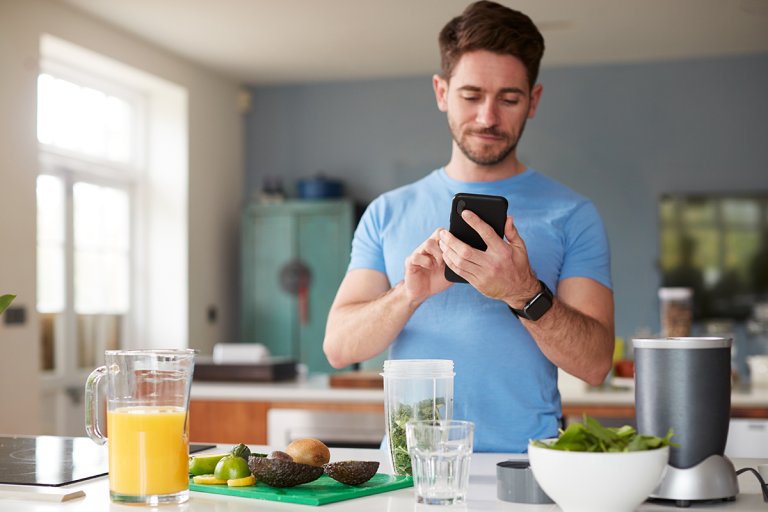 Man looking at his phone while making a smoothie in a kitchen.