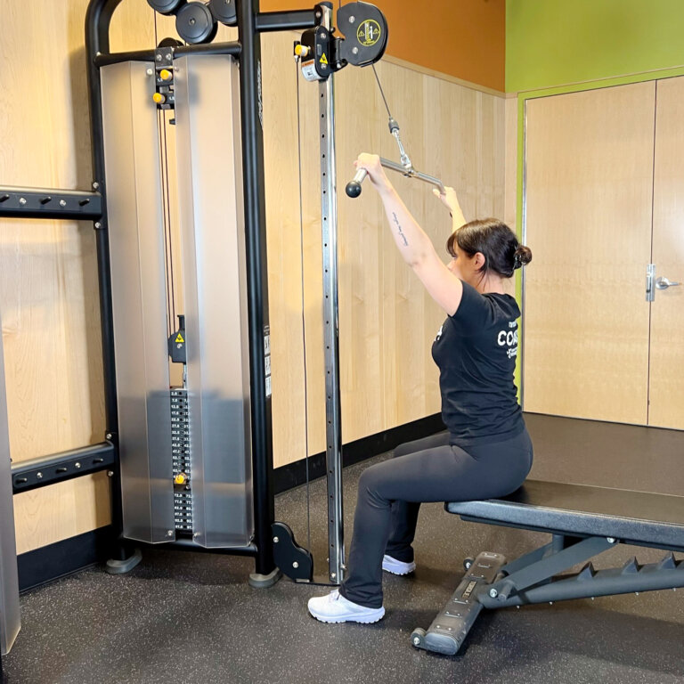 Coach seated on a bench, facing a cable machine, holding a lat bar.