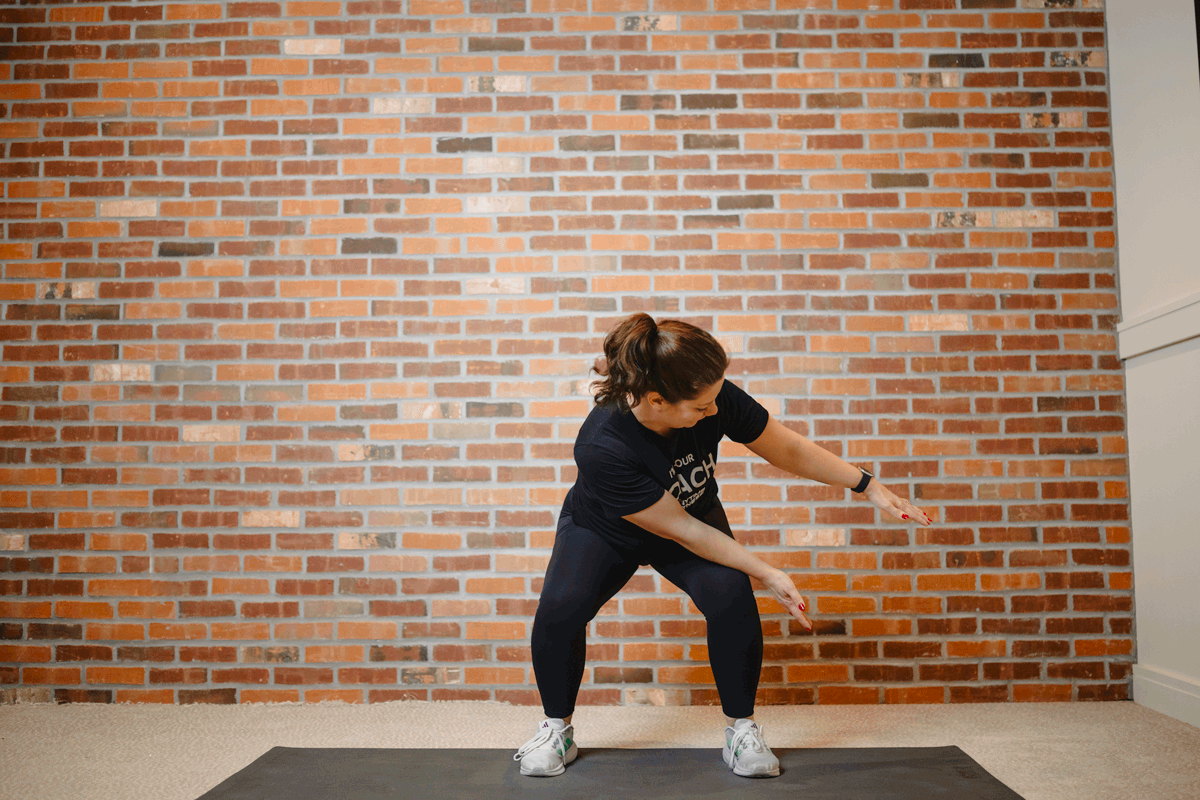 Anytime Fitness Coach performing air squats on an exercise mat in a home gym, targeting the upper and lower body simultaneously.