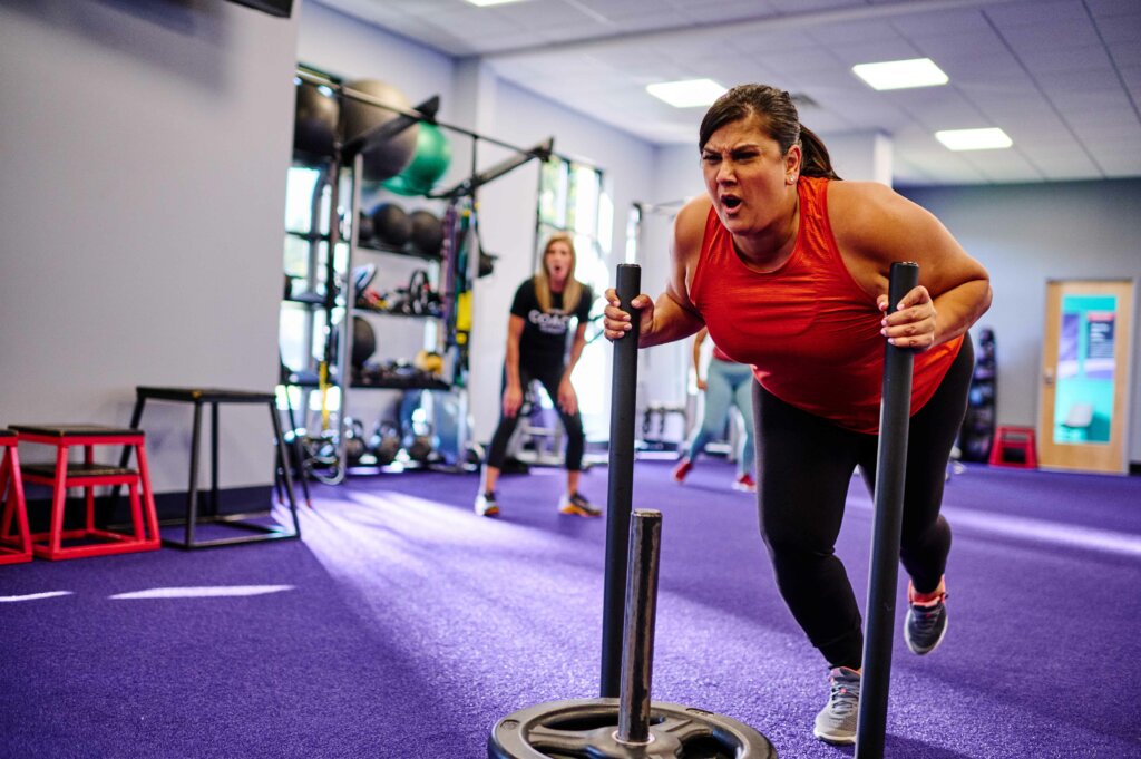 Woman doing a sled push with a coach cheering her on in the background.