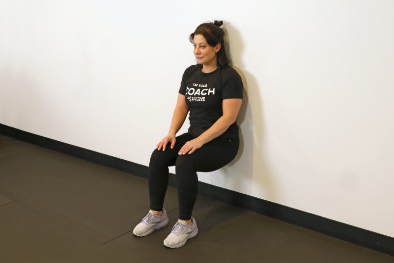 Coach Heather demonstrating a Wall Sit in a gym setting.