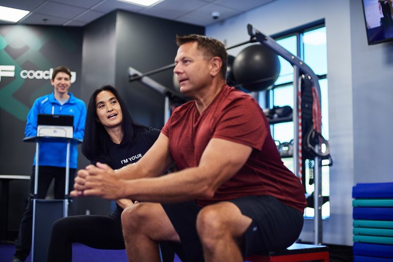 Man performing a squat in a gym with an Anytime Fitness Coach encouraging him.