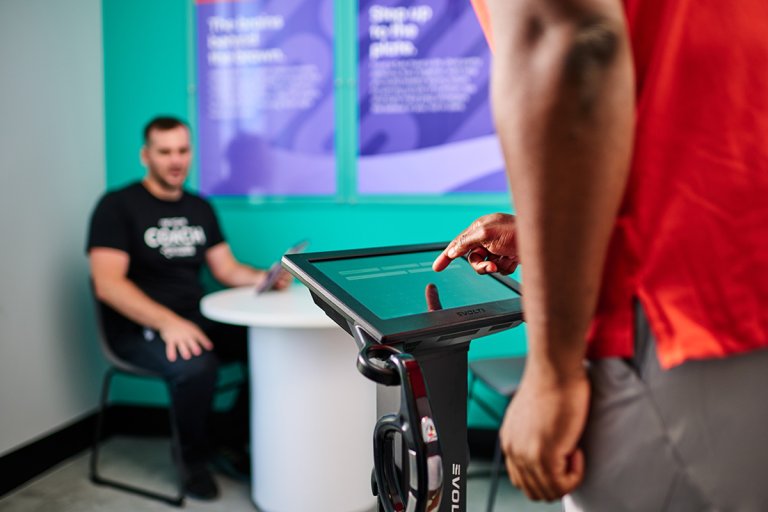 Man taps an Evolt 360 screen while an Anytime Fitness Coach looks on.