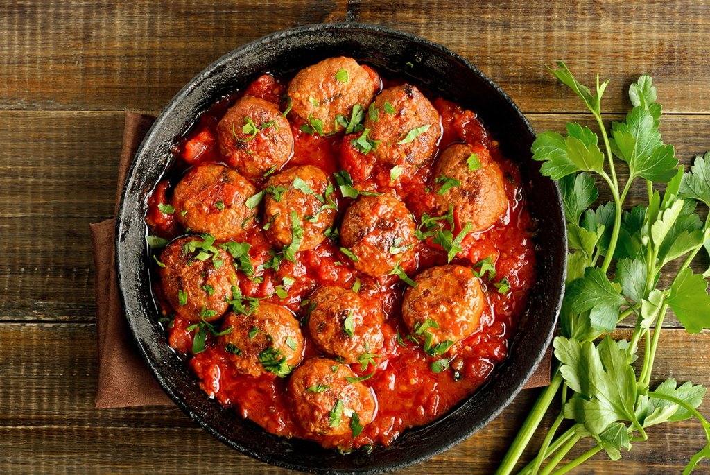Skillet of meatballs in red sauce with parsley sprigs on the table next to it.