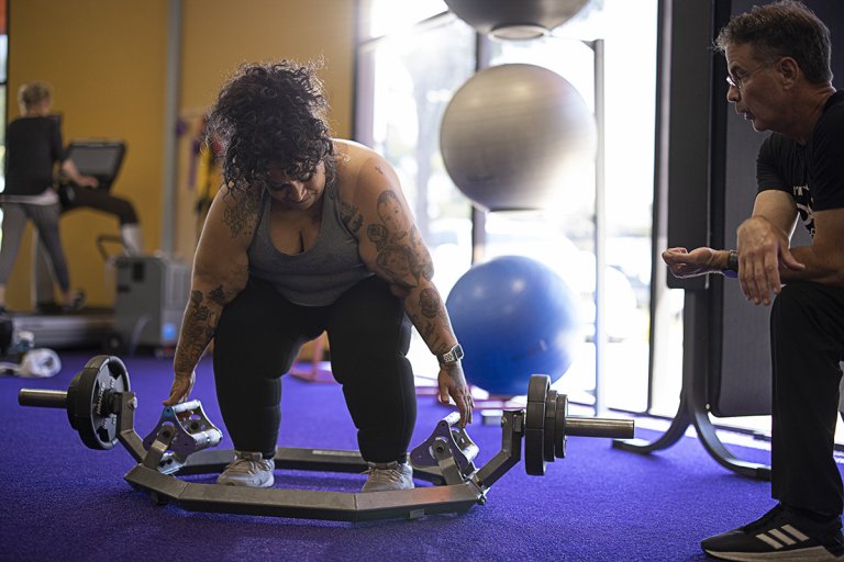 Candie bending down to lift a deadlift bar with her Coach, Mike, nearby in the gym offering support.