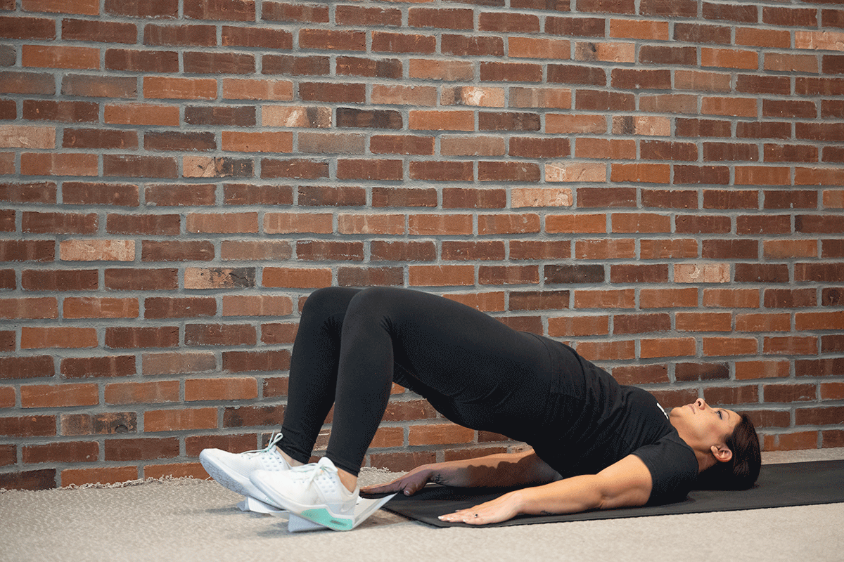Woman laying on her back on a yoga mat with her feet on sheets of paper, pulling her legs in and out.