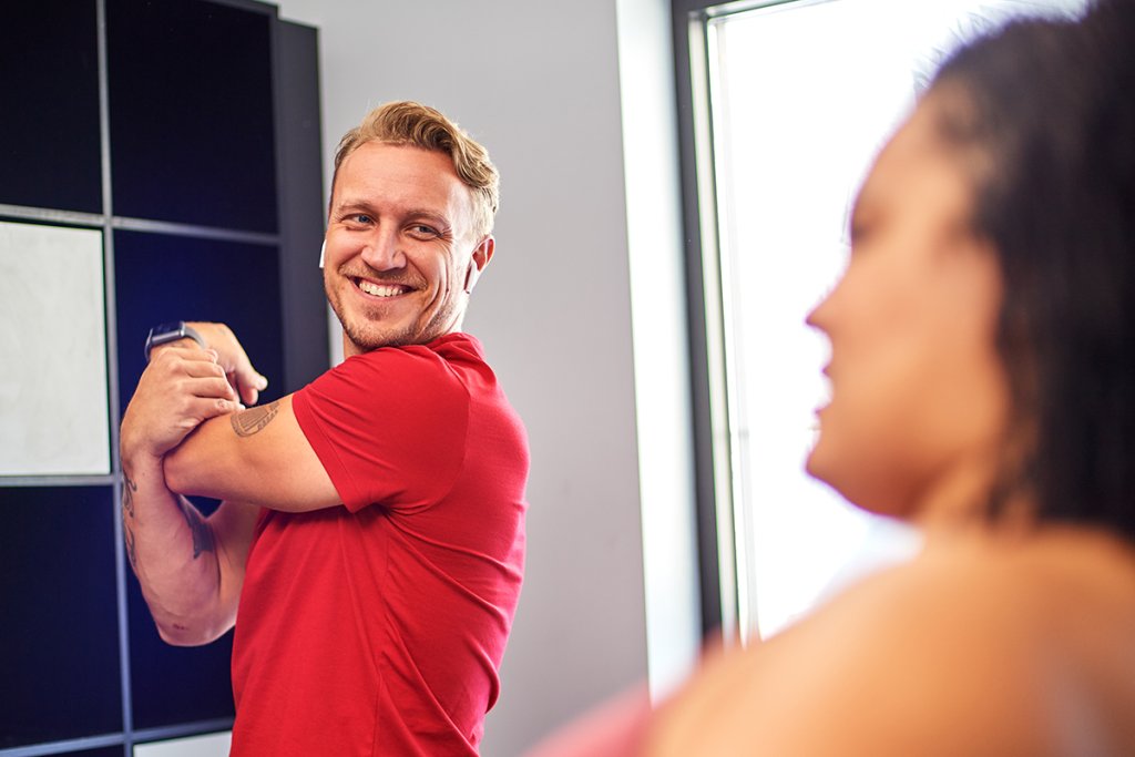 Man in a red shirt stretching in a gym locker area while talking to a woman.