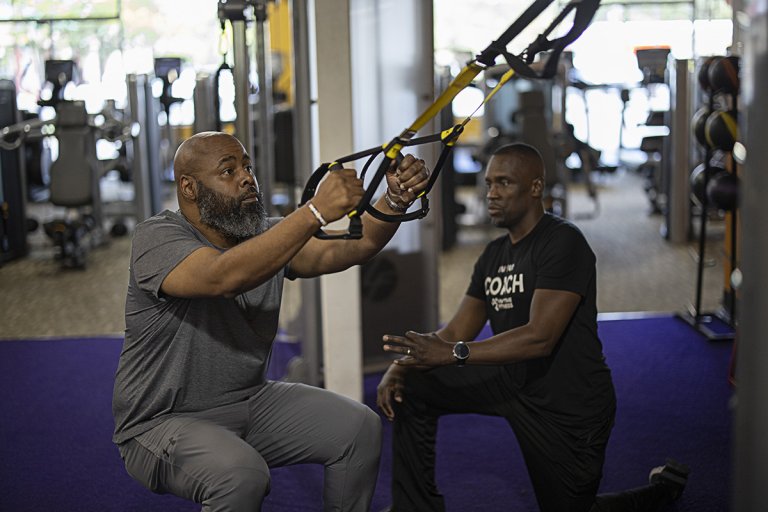 Coach Devon assists Mark with his form as Mark performs a TRX squat in a gym setting.