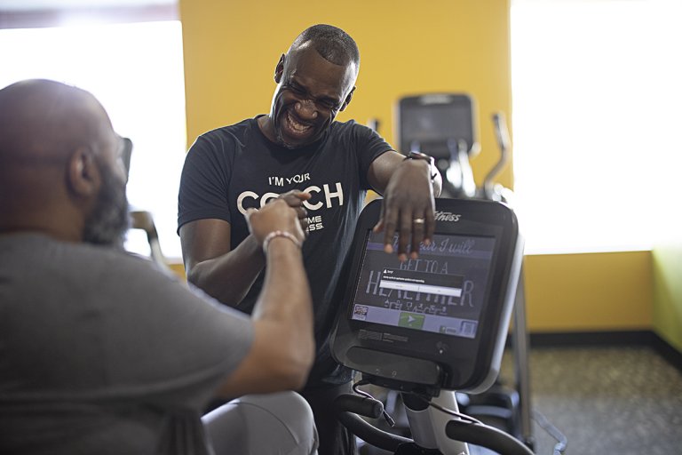 Coach Devon and Mark celebrate with a fist bump as Mark rides a recumbent bike in the gym.