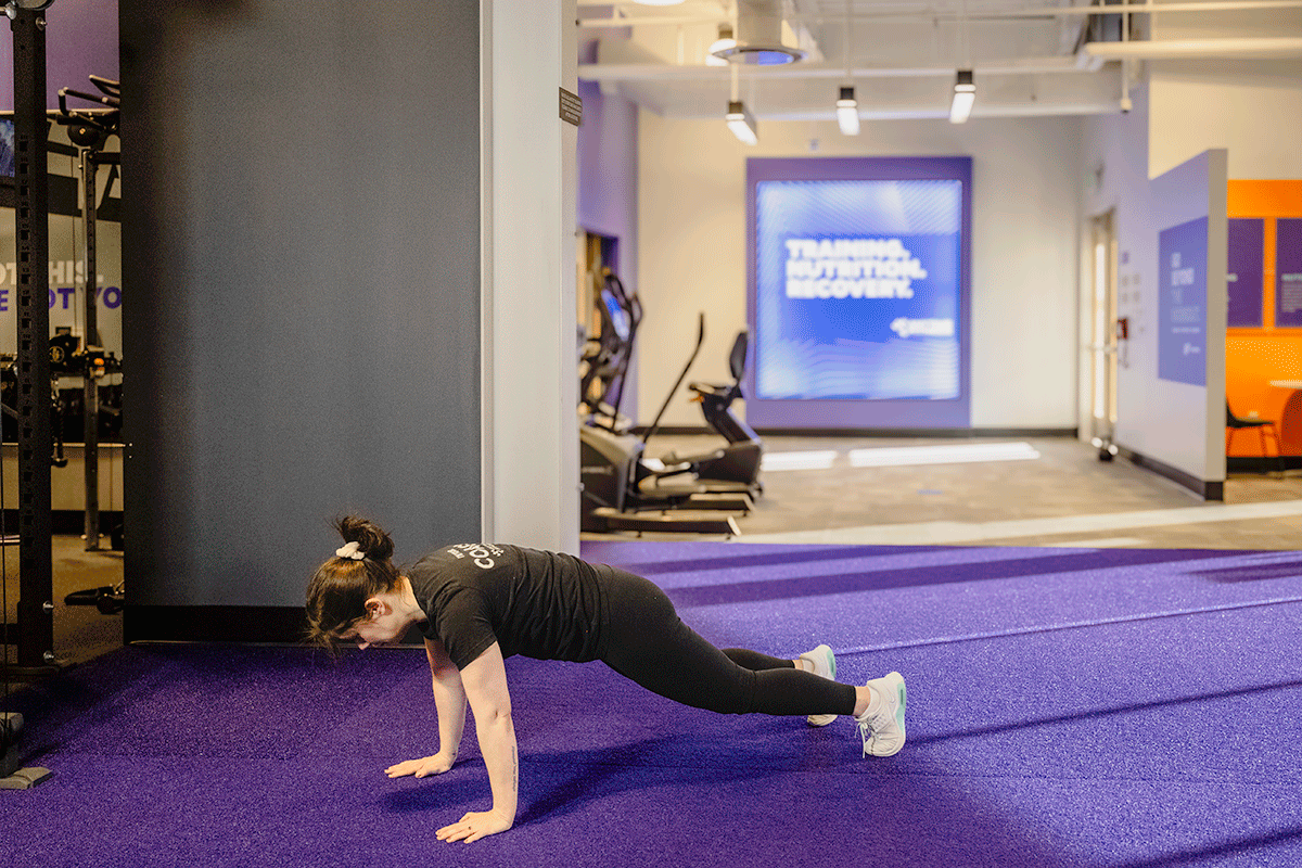 Woman in a gym squatting then jumping her feet back, doing a push-up, and jumping back to a standing position.