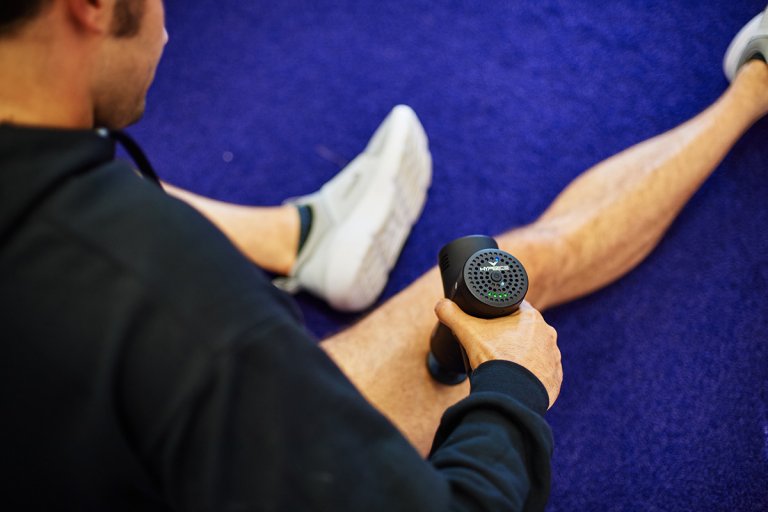 Man using a muscle massage percussion instrument on his thigh while sitting in a gym.