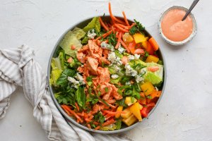Bowl of salad made with bright ingredients including romaine lettuce, cooked chicken, cubed bell peppers, shredded carrots, sliced green onions, parsley, and blue cheese, with a small bowl of creamy buffalo dressing on the side and a napkin nearby.