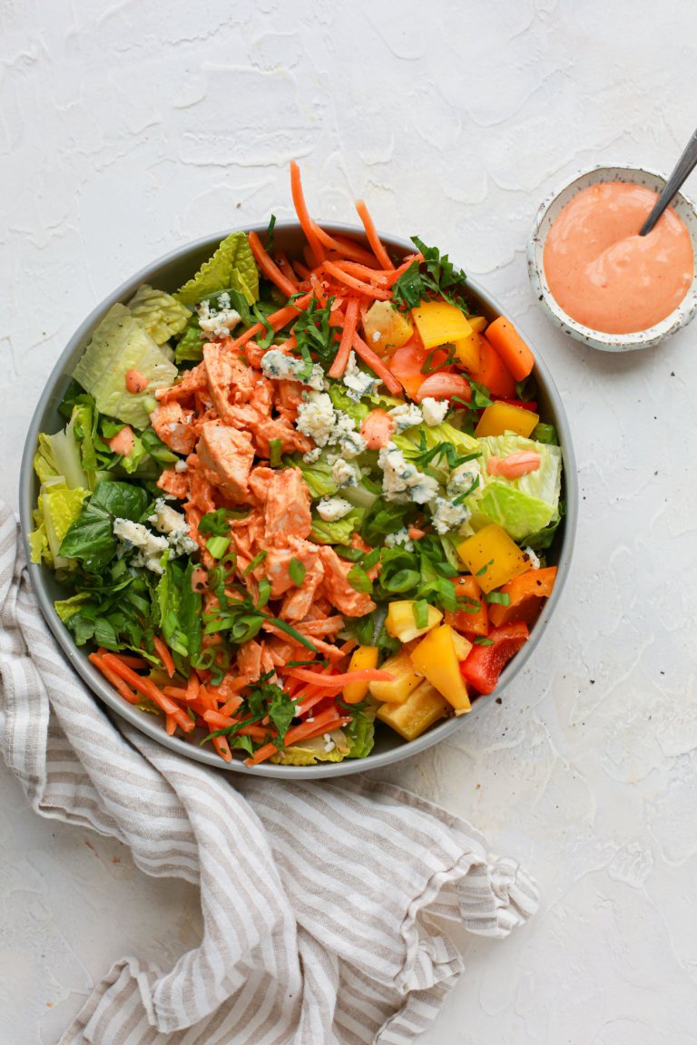 Bowl of salad made with bright ingredients including romaine lettuce, cooked chicken, cubed bell peppers, shredded carrots, sliced green onions, parsley, and blue cheese, with a small bowl of creamy buffalo dressing on the side and a napkin nearby.