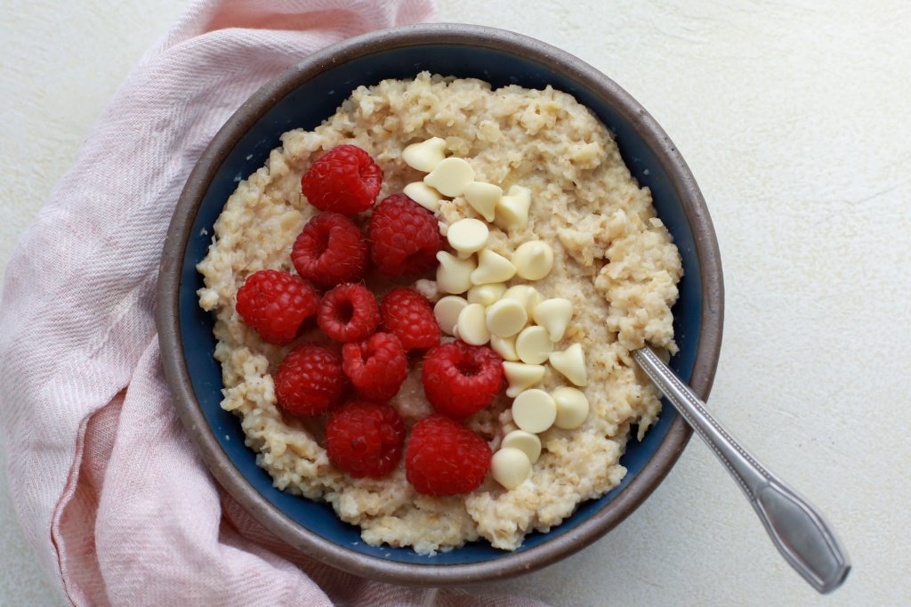 Bowl of oats prepared with raspberries and white chocolate chips as toppings. shown ready-to-eat with a spoon.
