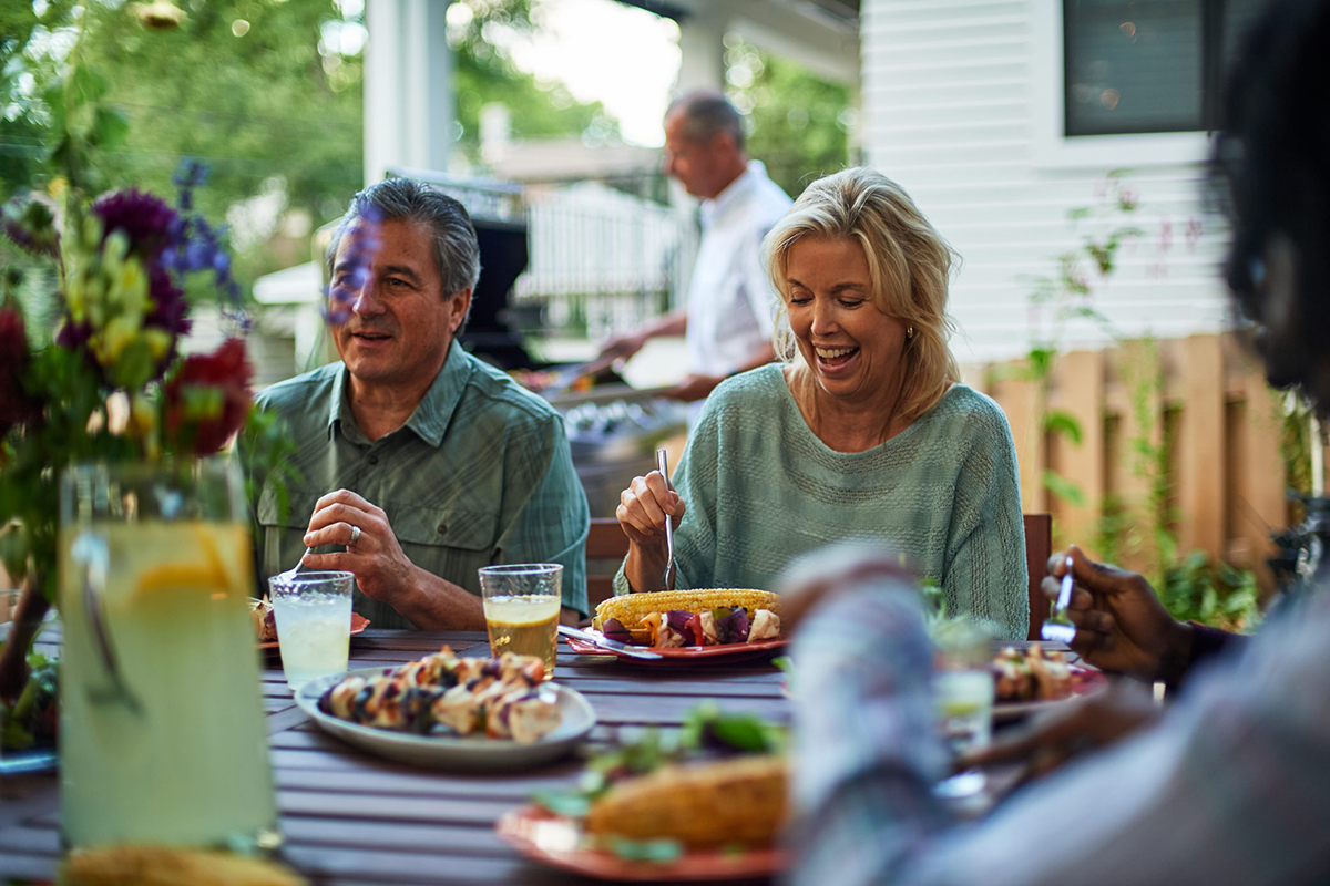 A group of middle-aged friends sit around an outdoor table eating grilled food in the summer.