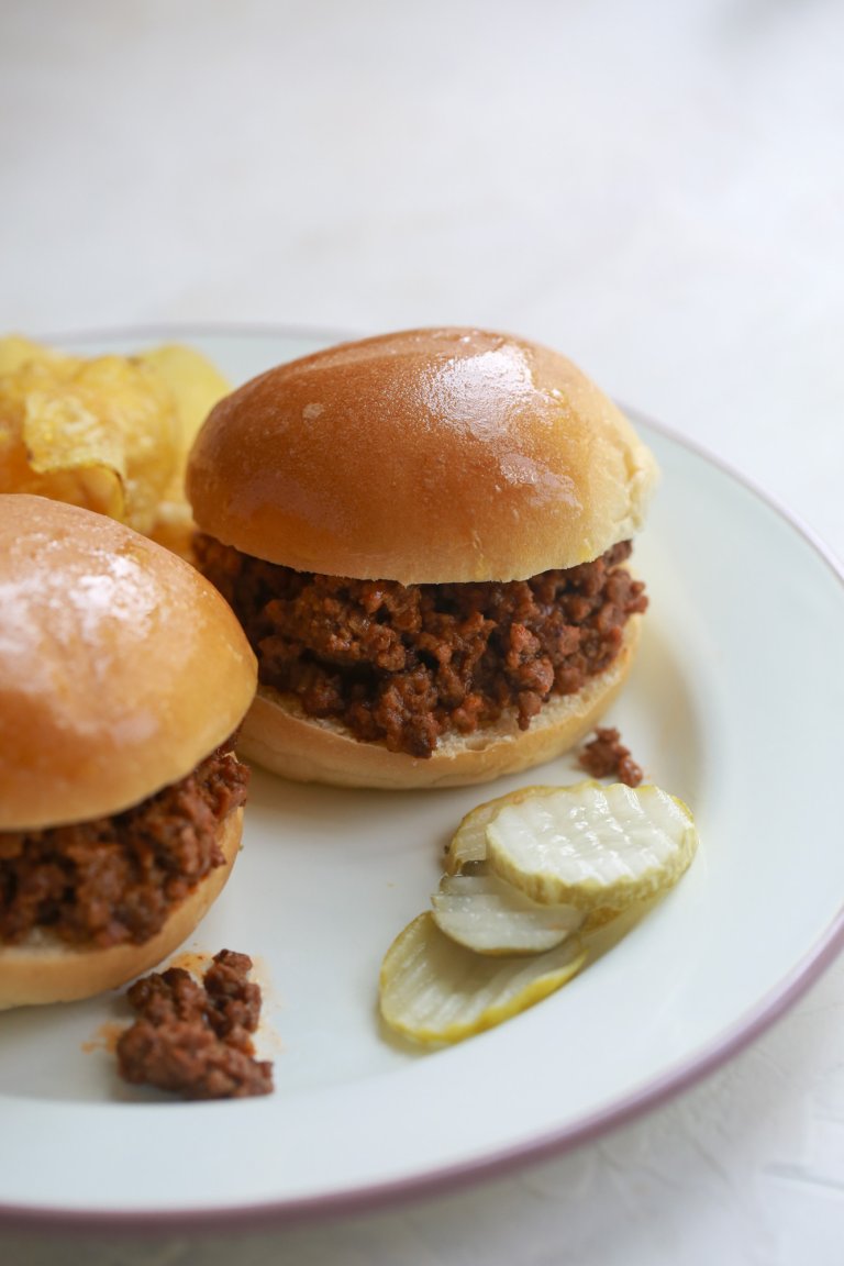 A plate with two Sloppy Joes sandwiches, potato chips and pickle slices.