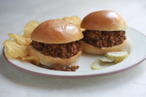 A plate with two Sloppy Joes sandwiches, potato chips and pickle slices.