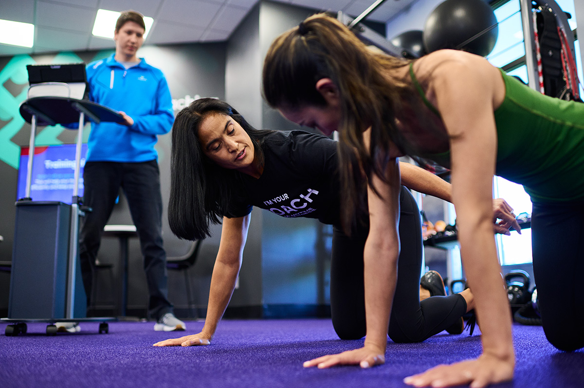 A fitness coach shows a client how to do a plank while a physical therapist looks on in the background.
