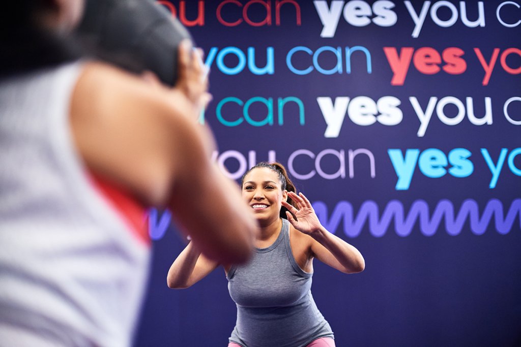 Smiling woman in workout gear engaging in a fitness class with motivational 'Yes You Can' background text.