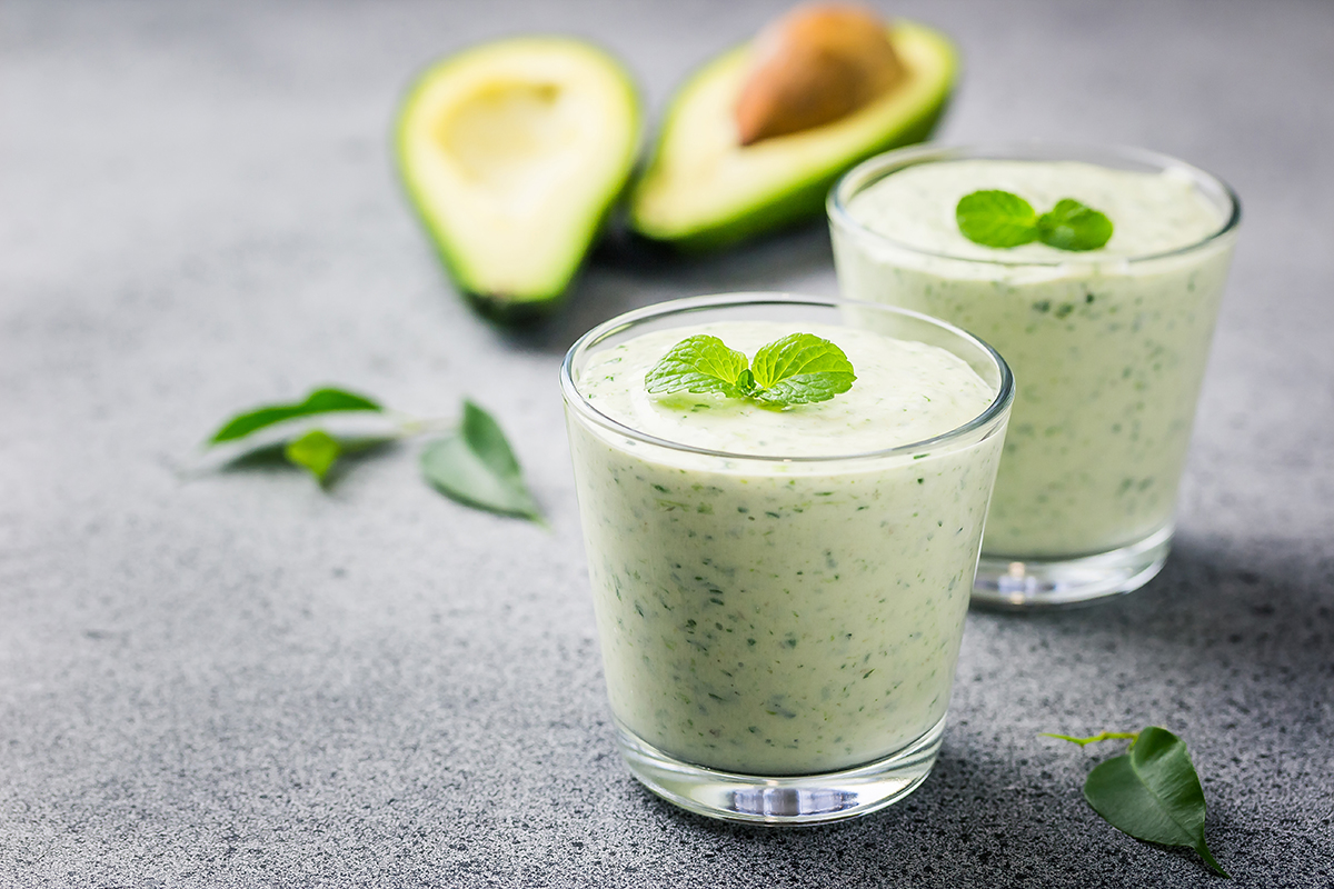 Two glasses of creamy avocado smoothies garnished with mint leaves, with fresh avocados in the background.