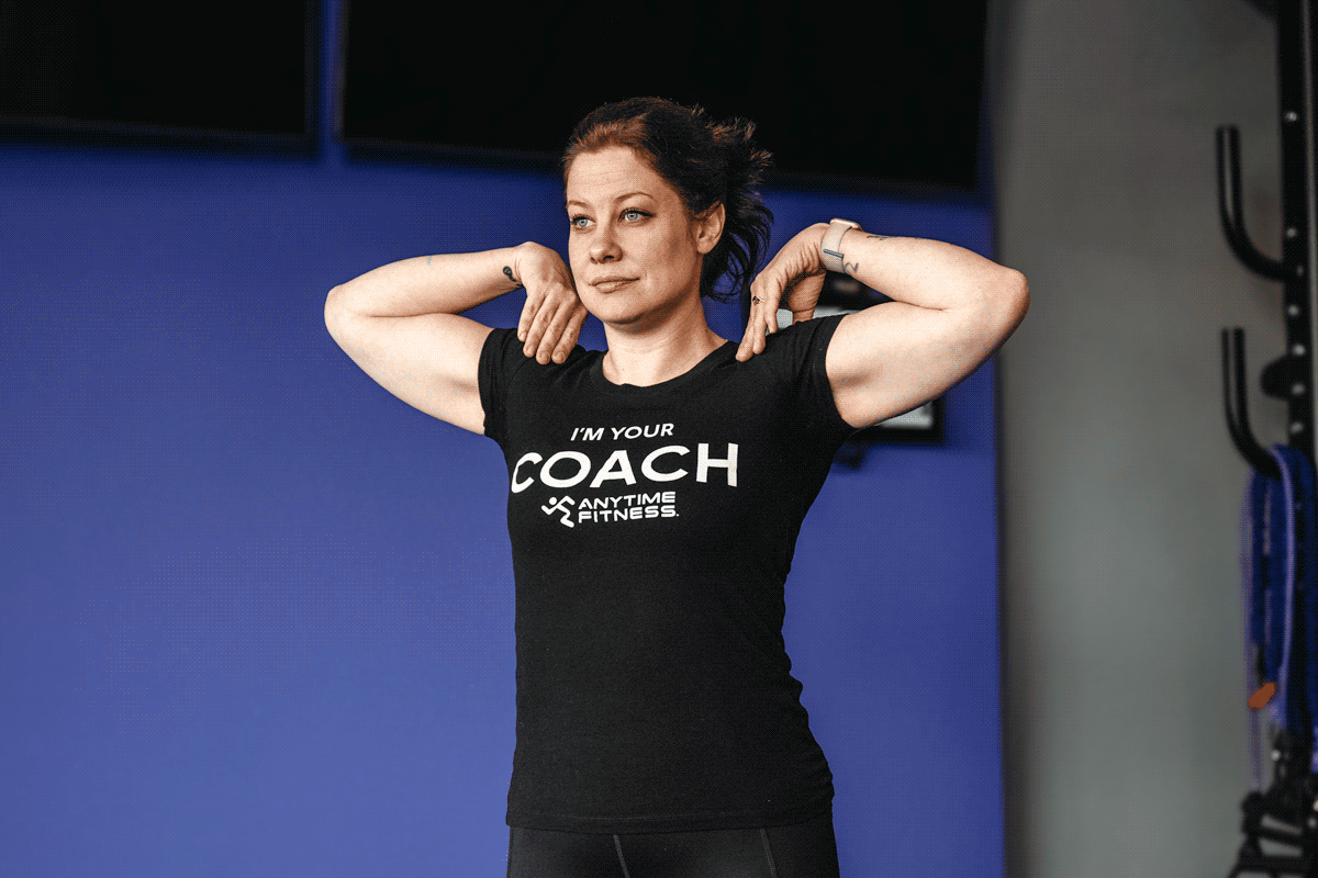 Anytime Fitness coach performing arm circles as part of a treadmill warmup routine, wearing a black 'I'm your coach' T-shirt.