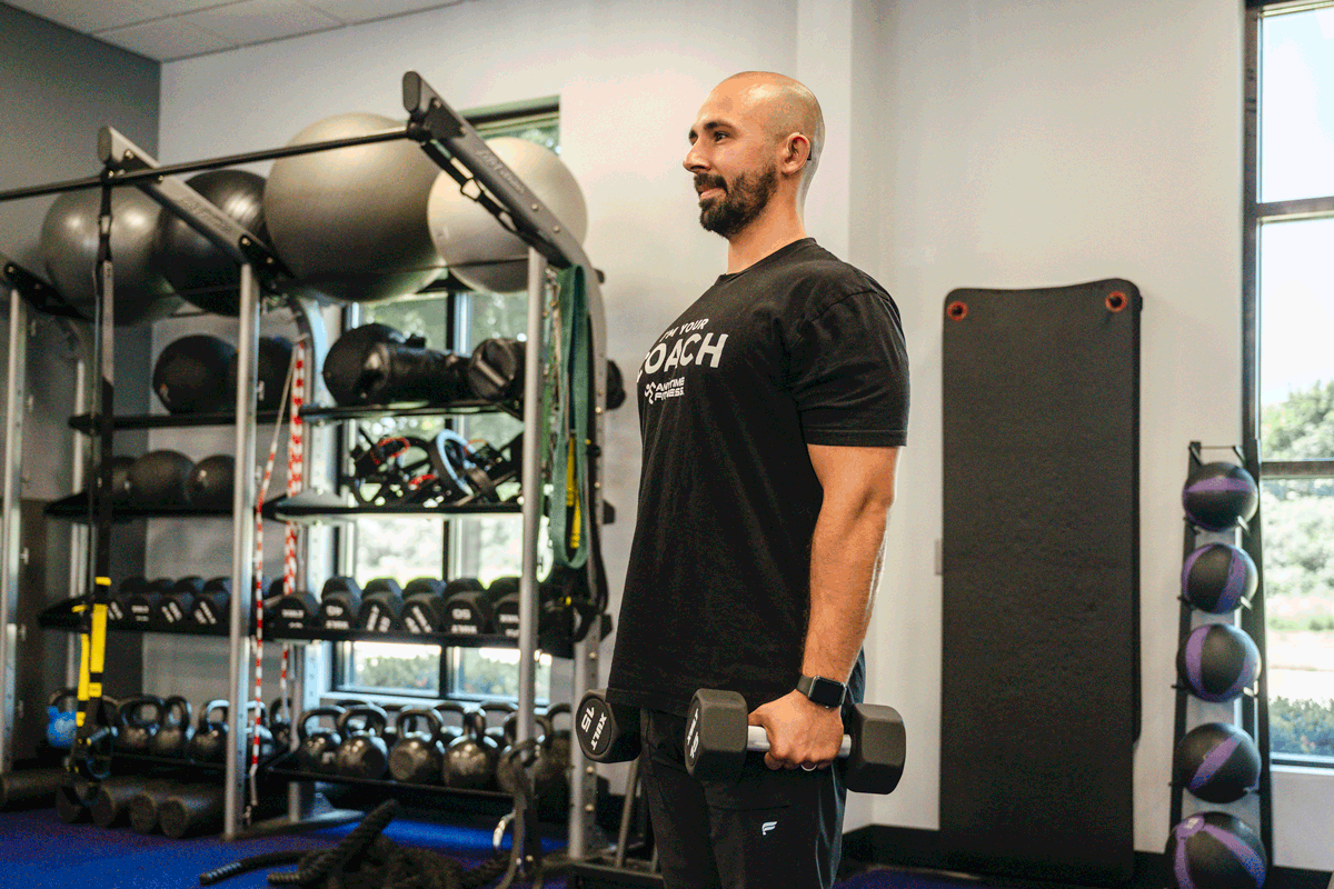 Anytime Fitness coach demonstrating front raise exercise with dumbbells, focusing on shoulder muscle engagement.