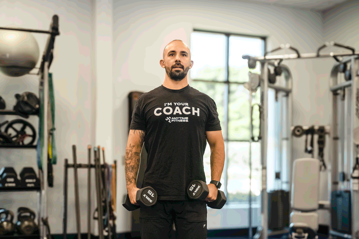 Anytime Fitness coach performing lateral raise exercise with dumbbells, highlighting shoulder muscle activation.