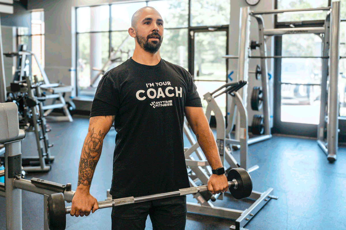 Anytime Fitness coach performing upright row exercise with a barbell, showcasing proper shoulder workout form in a gym setting.