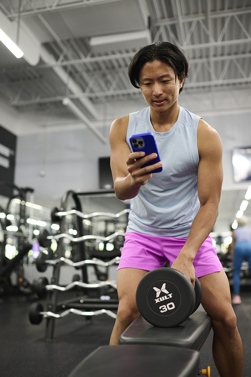 Person in a gym holding a phone, preparing to do a strength workout with a dumbbell.