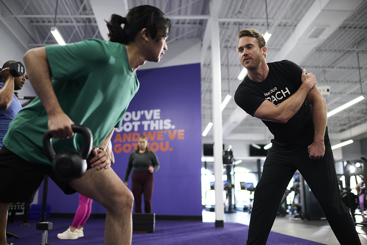 A gym member is working with an Anytime Fitness Coach. The Coach is demonstrating how to perform an exercise while the member holds a weight in his right hand.