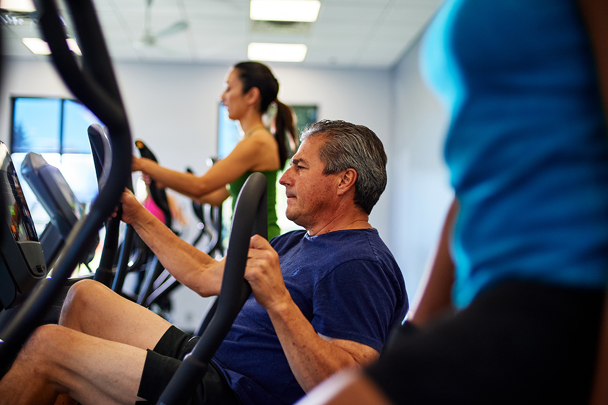Several gym members are shown exercising on various machines, including the elliptical and bike. A man is in the middle of the row of people using exercise equipment and is focused on his workout.