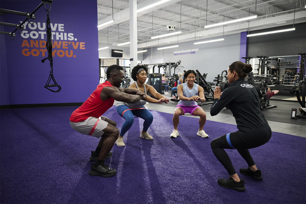 An Anytime Fitness Coach leads a training session for a group performing squats with resistance bands.