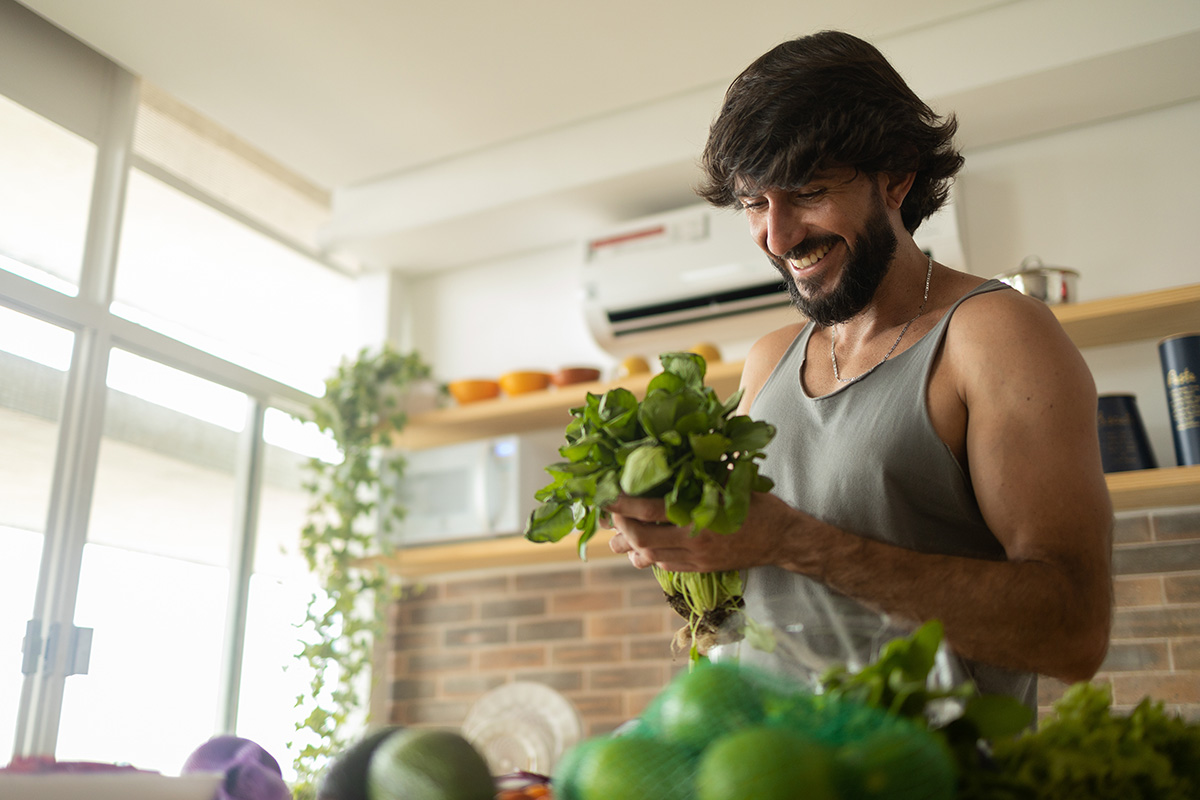 Man in a gray tank top sorting through a variety of green produce, including herbs, lettuce, and limes.
