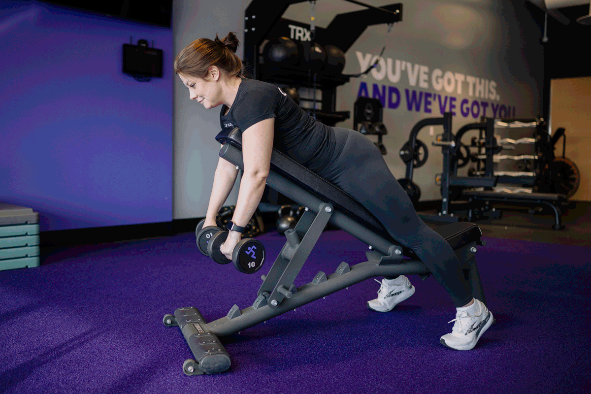An Anytime Fitness Coach demonstrating a chest-supported incline row on a bench with dumbbells.